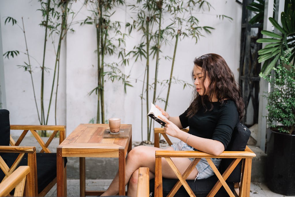 Woman reading a book, sitting in an outdoor café surrounded by lush greenery.