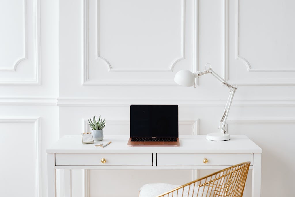 Elegant minimalist home office desk setup with a laptop, lamp, and indoor plant in a bright white space.